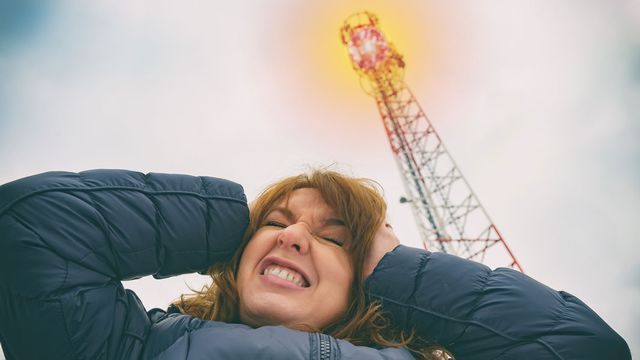 Une femme se tient la tête près d'une antenne émettrice de réseaux cellulaires. [Amaviael - Depositphotos]