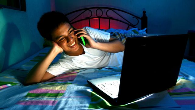 Un adolescent discute au téléphone tout en regardant son ordinateur. [Junpinzon - Depositphotos]