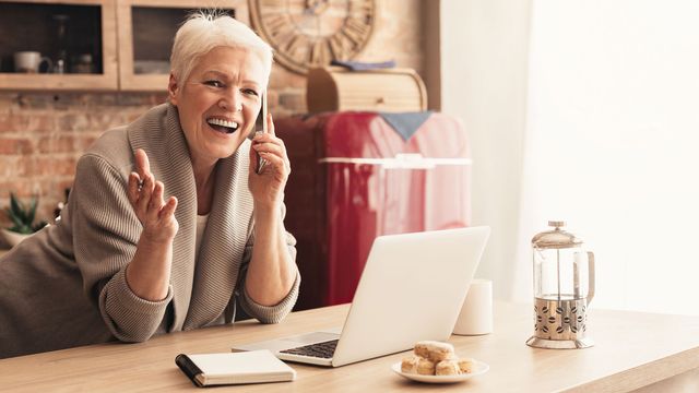 Une femme senior parle au téléphone en consultant son ordinateur portable. [Milkos - Depositphotos]