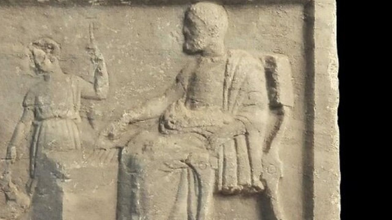 La scène représente un homme barbu et un garçon positionnés autour d’un ensemble de cinq lignes gravées sur un bloc [Courtesy Larissa Museum - Diachronic Museum of Larissa]