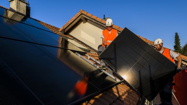 Deux installateurs implantent des panneaux solaires sur le toit d'une maison. [Jean-Christophe Bott - Keystone]